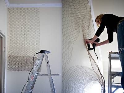 A woman is stapling strips of chicken wire onto wall.