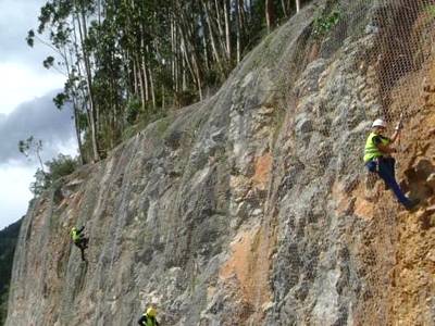 Four workers are fixing hexagonal wire netting rockfall barrier to a side of mountain near the road